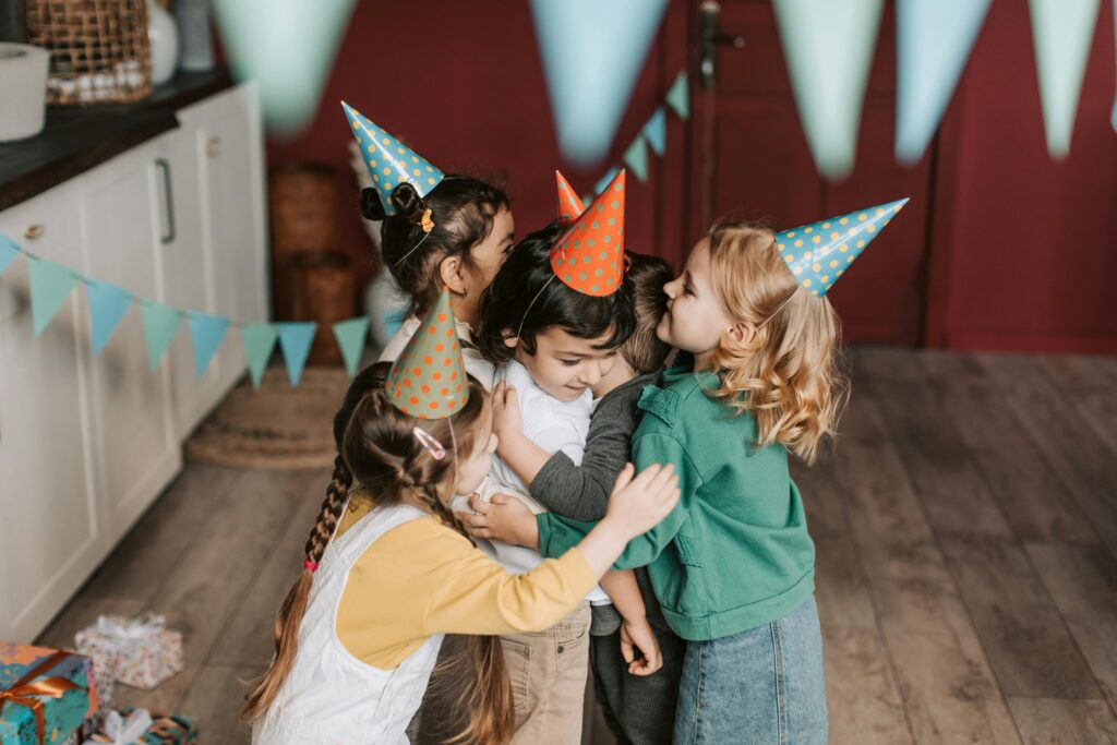 Children wearing party hats embrace each other during a joyful indoor birthday celebration.